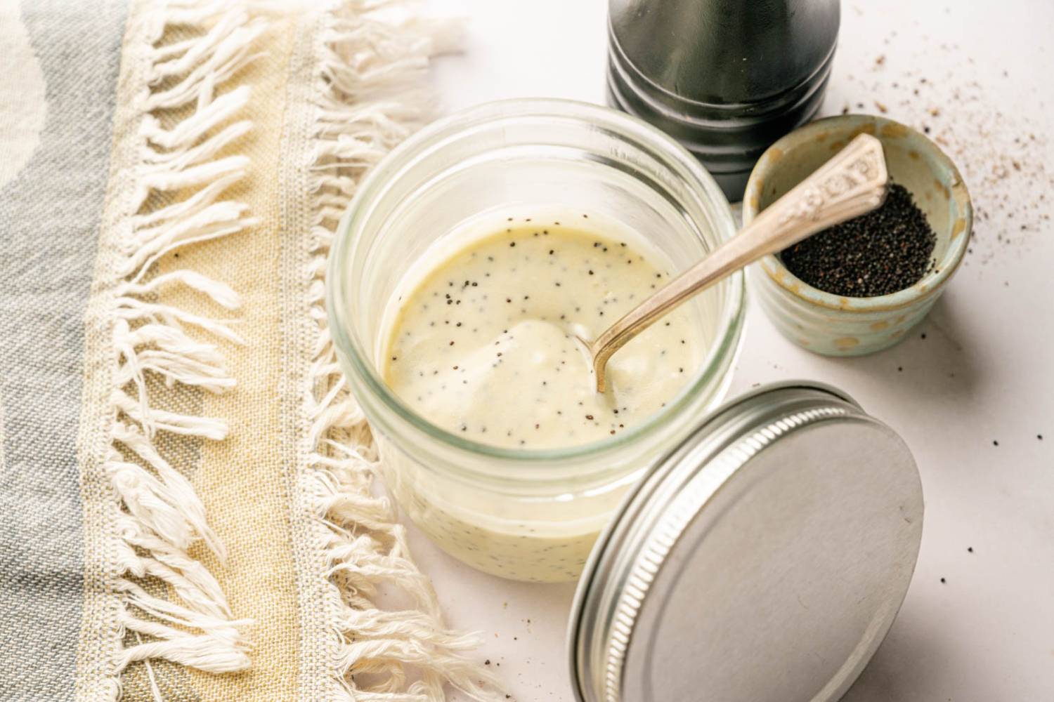 Jar of healthy poppy seed dressing with black pepper and poppy seeds nearby, styled on a neutral kitchen surface with a linen towel.