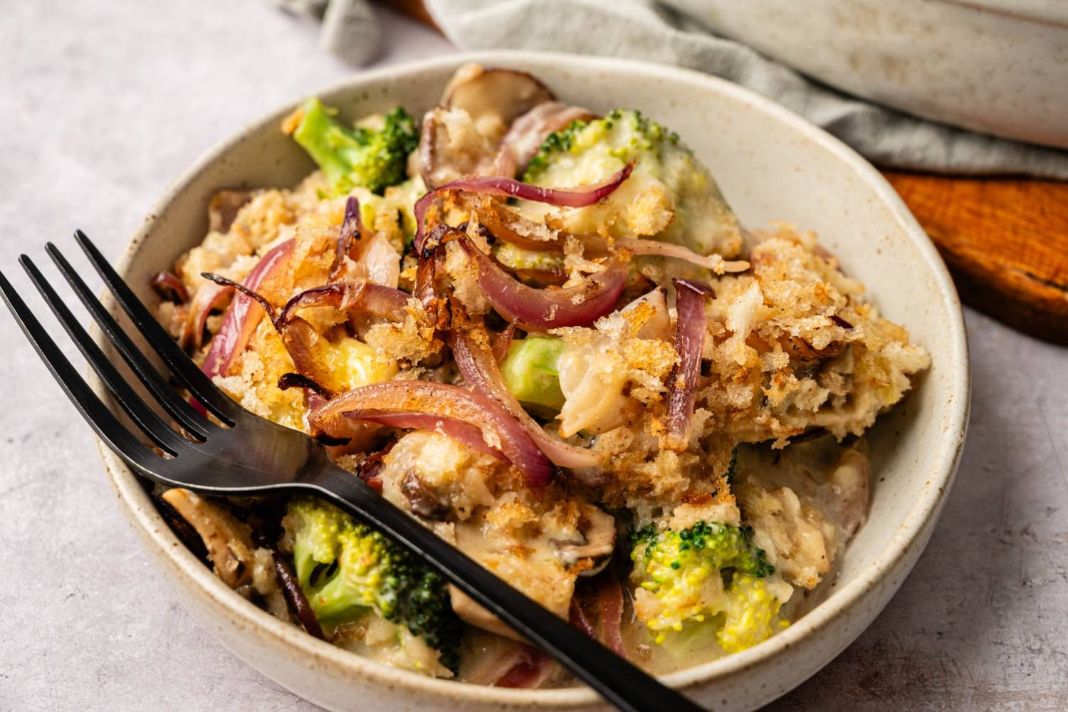 Healthy Broccoli Casserole A bowl of broccoli and mushroom casserole topped with crispy breadcrumbs and caramelized onions. A fork rests on the bowl, adding a cozy, inviting feel.
