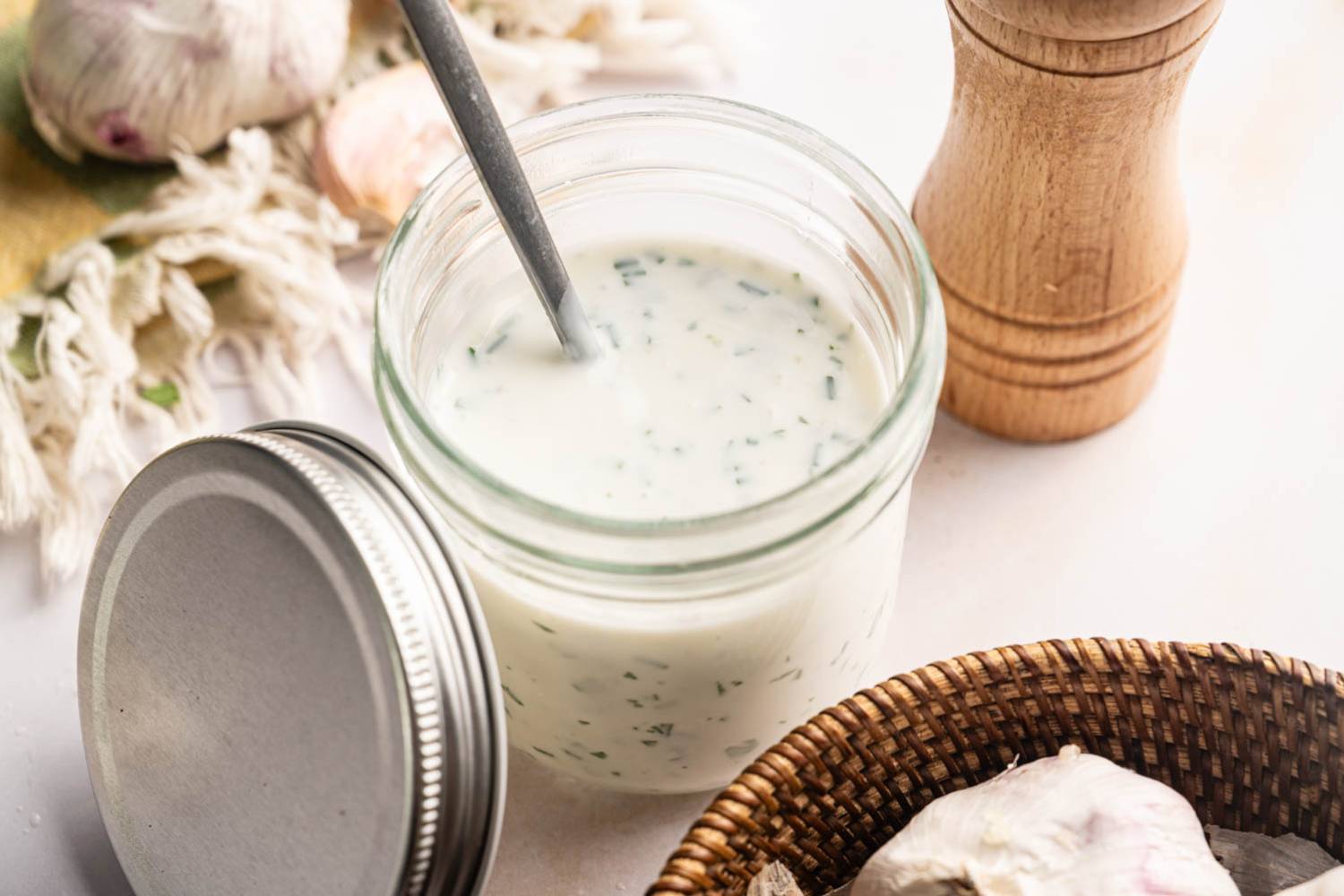 Close-up of ranch dressing in a glass jar with a spoon stirring the mixture, the lid resting to the side, and a wooden pepper mill and garlic bulbs in the background.