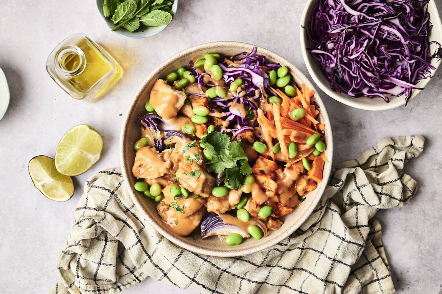 A vibrant bowl of chicken, edamame, purple cabbage, and carrots drizzled with sauce, surrounded by lime, mint, and olive oil.