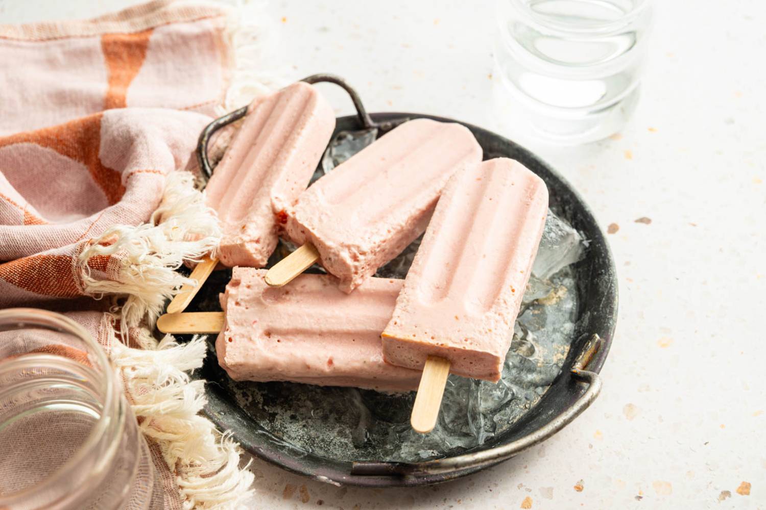 High Protein Greek Yogurt Popsicles Four homemade high-protein Greek yogurt popsicles resting on ice in a metal serving tray with a striped kitchen towel nearby.