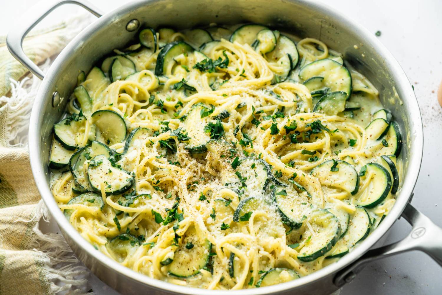 Close-up angled shot of creamy zucchini pasta in a stainless steel skillet, topped with Parmesan and herbs, styled on a marble countertop with a folded green and white linen napkin in the background.
