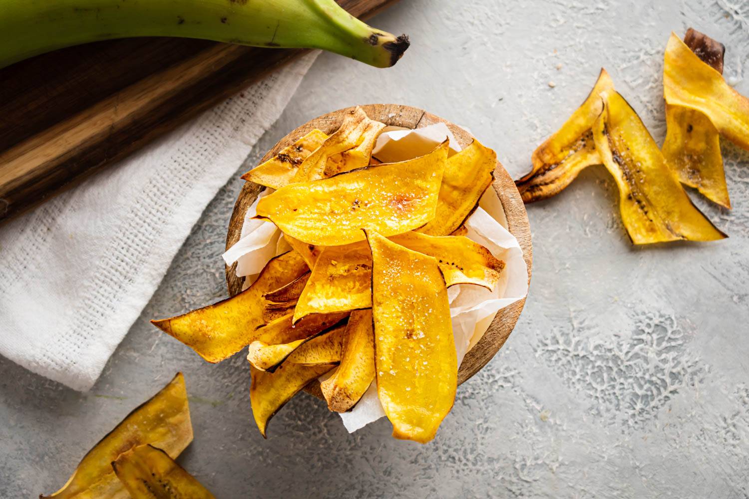 Homemade Plantain Chips Wooden bowl filled with golden homemade plantain chips, sprinkled with salt, surrounded by extra chips and a whole green plantain on a textured gray surface.