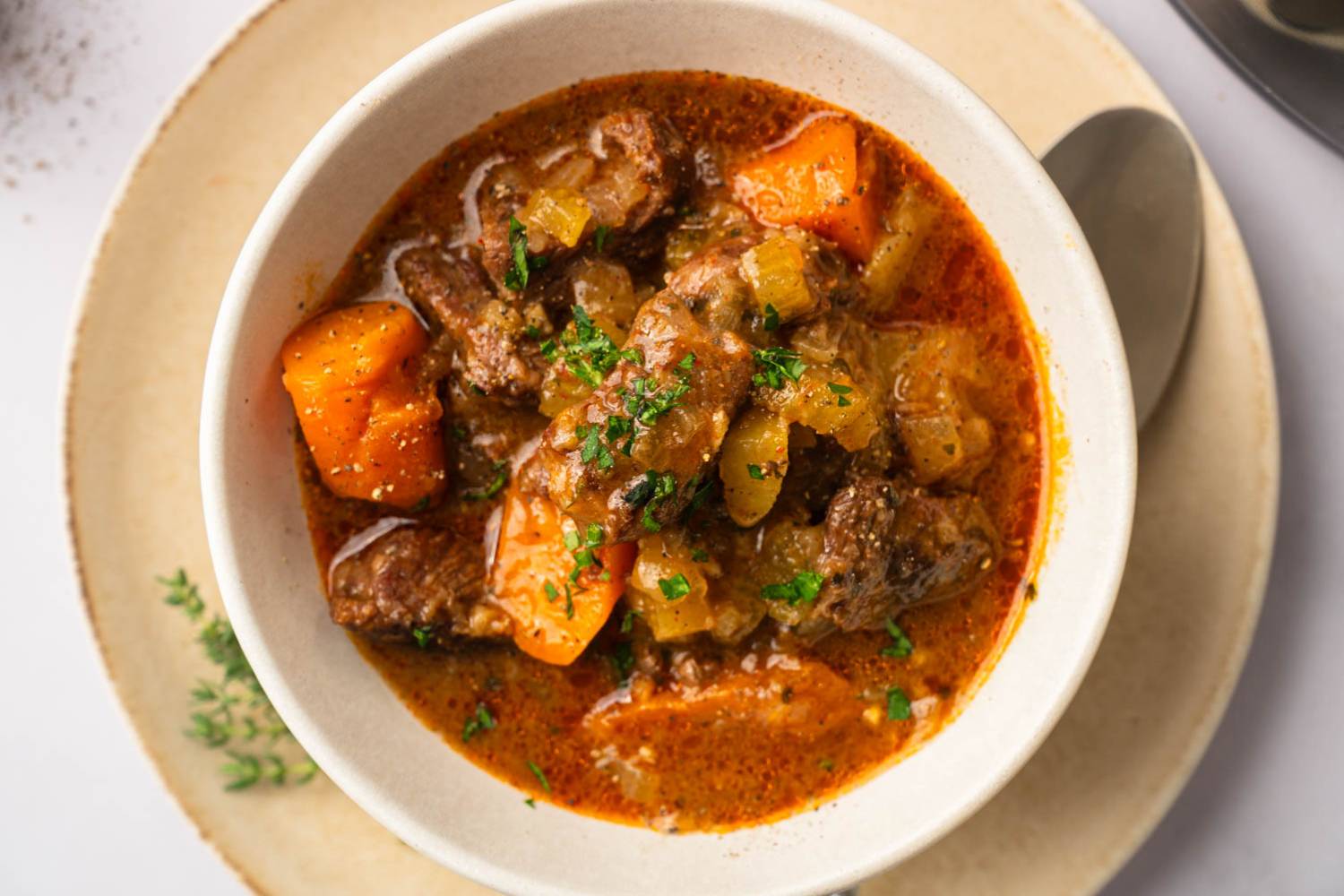 Healthy Instant Pot Beef Stew (or Slow Cooker) A bowl of hearty beef stew with chunks of carrot, celery, and garnished with fresh parsley, served on a cream-colored plate.