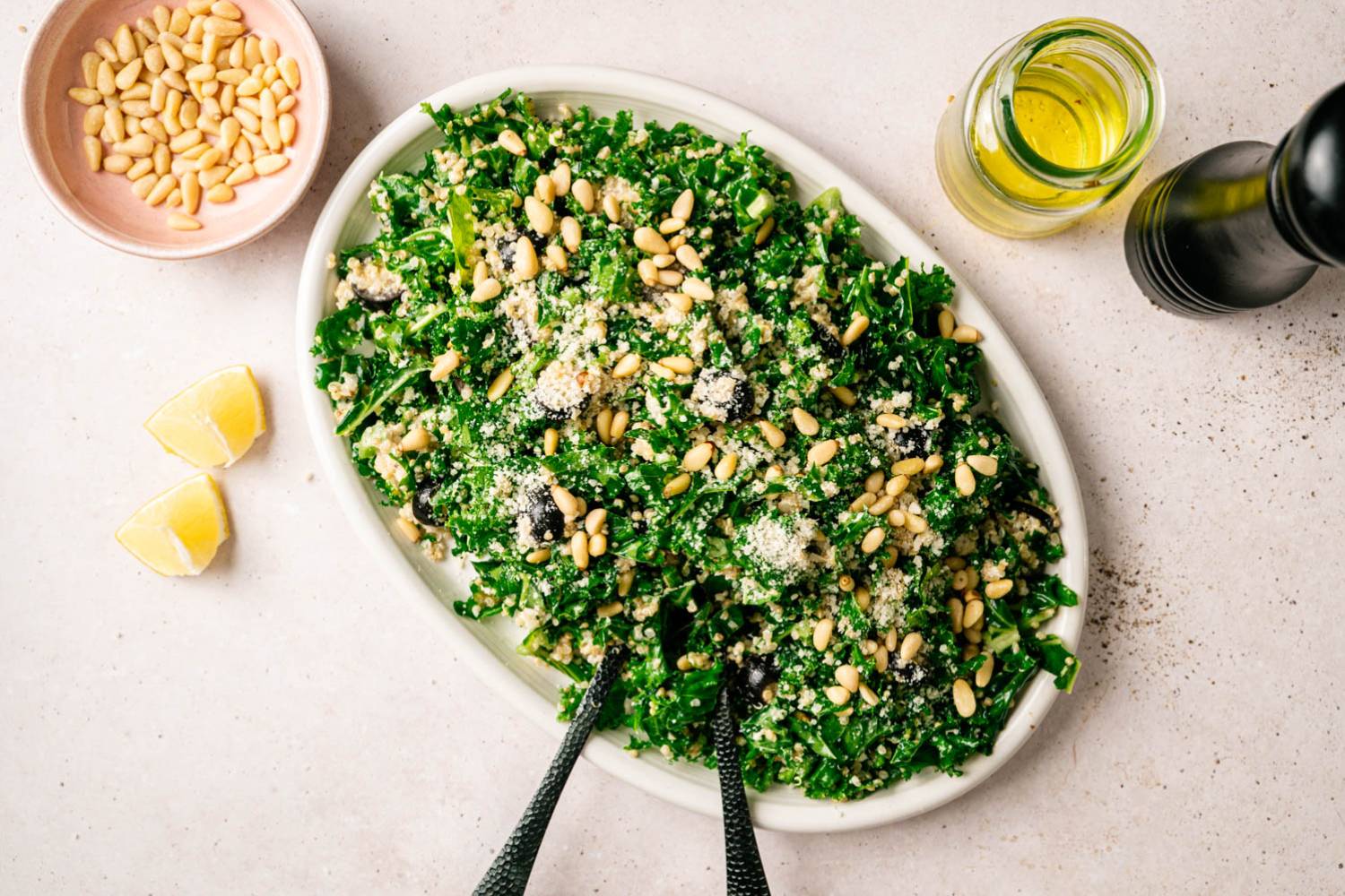 A vibrant kale salad on a white oval plate, garnished with pine nuts, grated cheese, and olives. Nearby are lemon wedges, olive oil, and a pepper grinder.