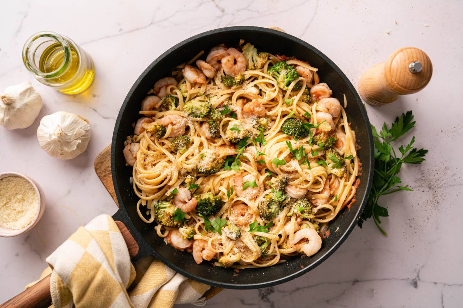 A skillet of shrimp and broccoli linguine, garnished with parsley and Parmesan. Surrounding are garlic cloves, olive oil, and a pepper mill on a marble surface.