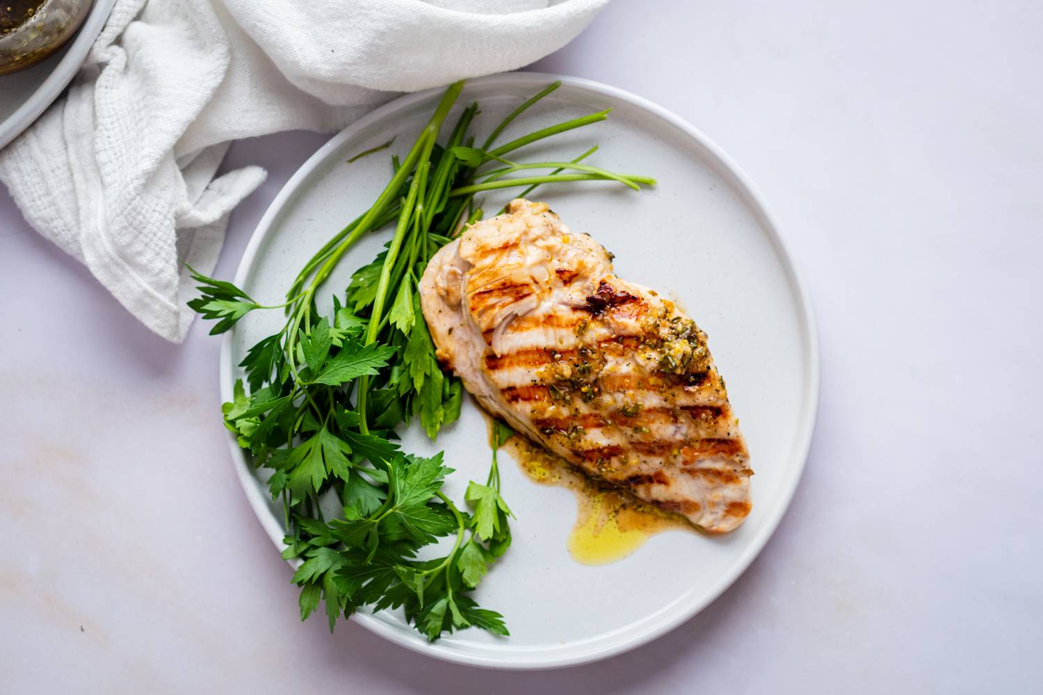 Lemon Pepper Marinade Grilled chicken breast with herb seasoning, served on a white plate with fresh parsley. Simple and appetizing presentation on a light background.