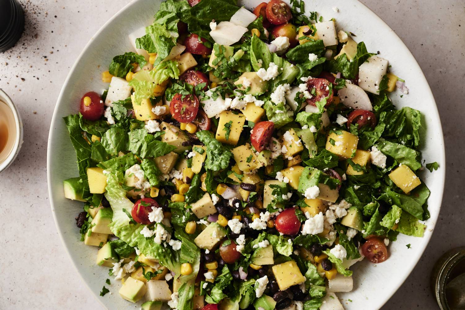 Mexican Chopped Salad with Honey Lime Dressing A vibrant Mexican chopped salad filled with diced mango, avocado, cherry tomatoes, black beans, corn, jicama, romaine lettuce, and crumbled cheese, garnished with fresh cilantro on a white plate.