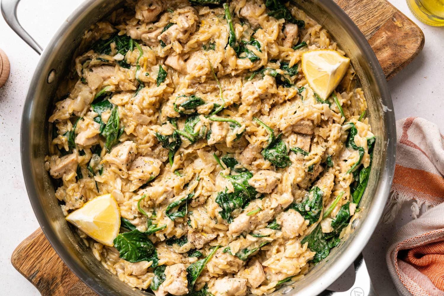 Top down shot of creamy chicken orzo with spinach and lemon wedges in a skillet set on a wooden board beside a pepper mill and orange striped towel.