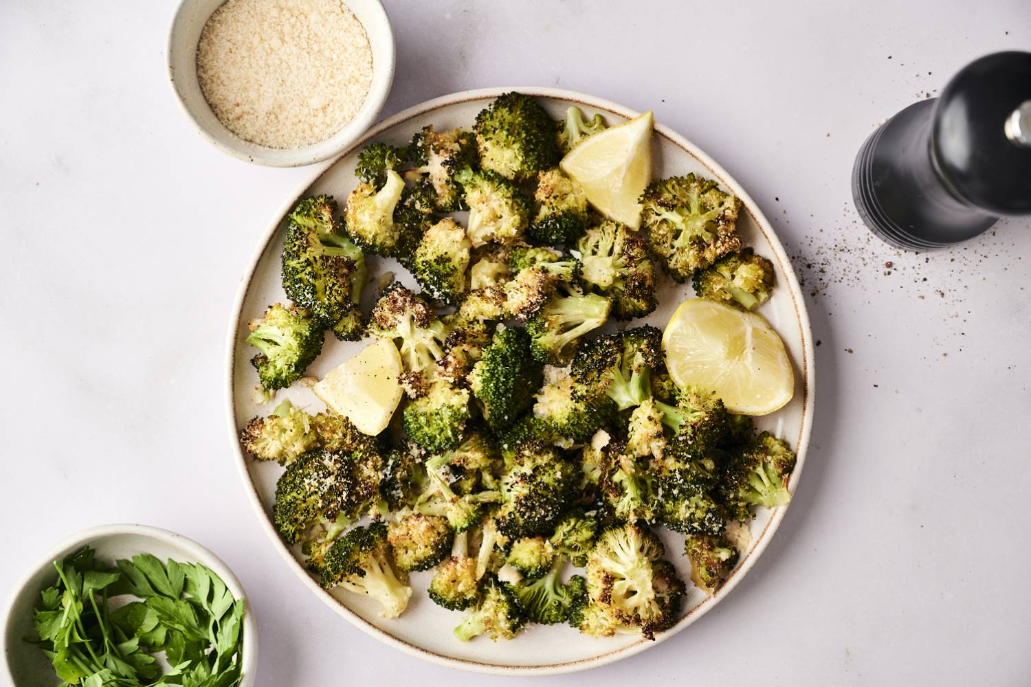 Parmesan Roasted Broccoli A plate of roasted broccoli garnished with lemon wedges, surrounded by parsley, grated cheese, and a pepper grinder.