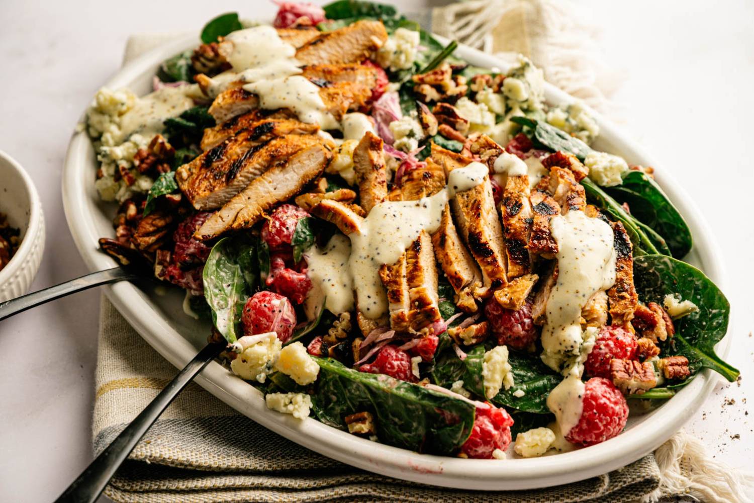  vibrant salad on a white platter featuring grilled chicken strips, fresh raspberries, spinach, pecans, blue cheese crumbles, and creamy dressing.