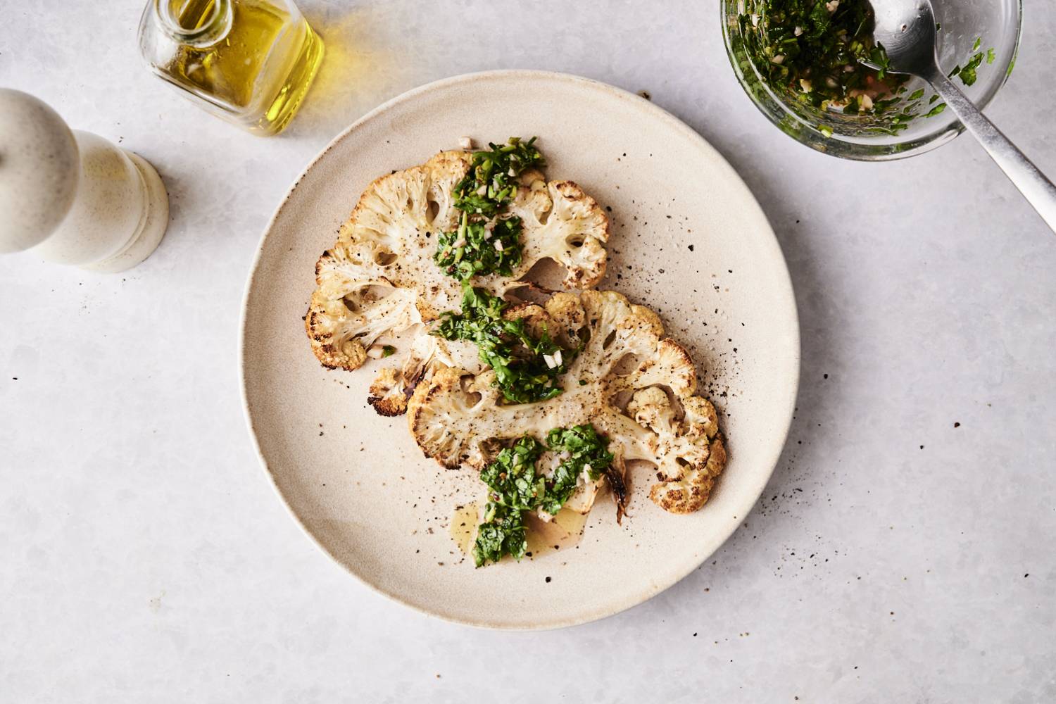 Cauliflower Steaks Overhead view of roasted cauliflower steaks garnished with chimichurri sauce, served on a plate with olive oil and pepper mill nearby.