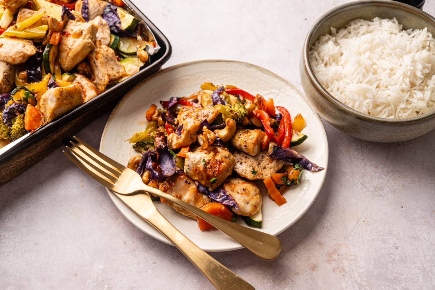 Stir-fried chicken with colorful vegetables, including red peppers and purple cabbage, on a white plate next to a bowl of white rice.