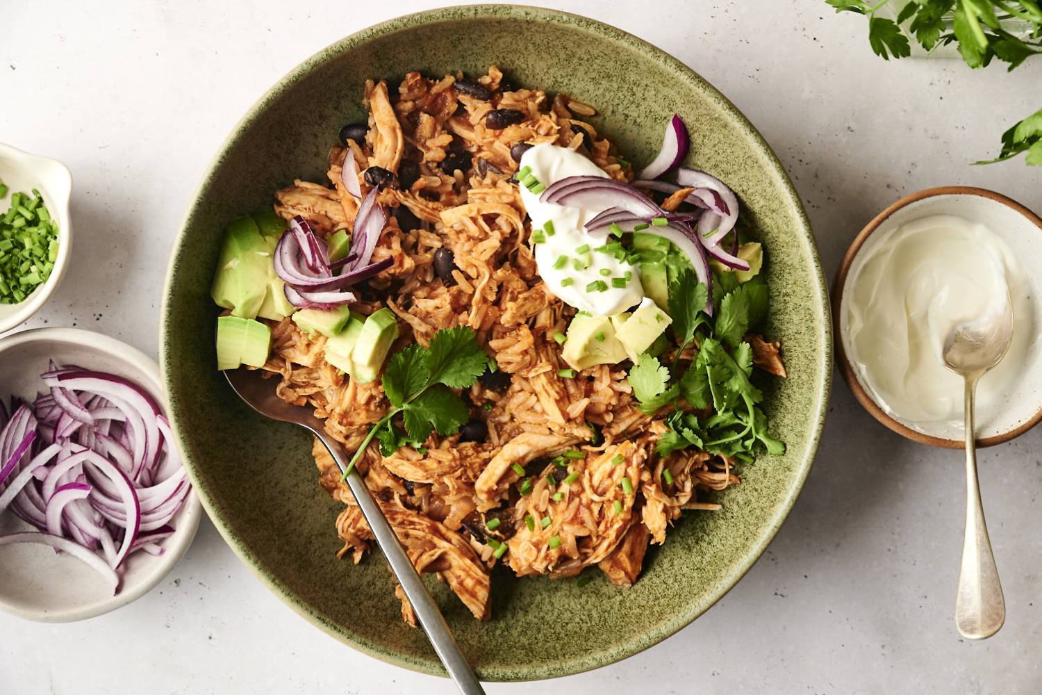 Chicken burrito bowl made with shredded chicken, rice, and black beans, topped with avocado, sliced red onion, sour cream, fresh cilantro, and chives. Small bowls of sour cream and red onion are arranged alongside the dish.