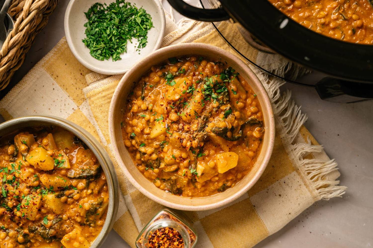 Overhead view of two bowls of creamy slow cooker Madras lentils with potatoes and spinach, garnished with fresh parsley and served beside a slow cooker.
