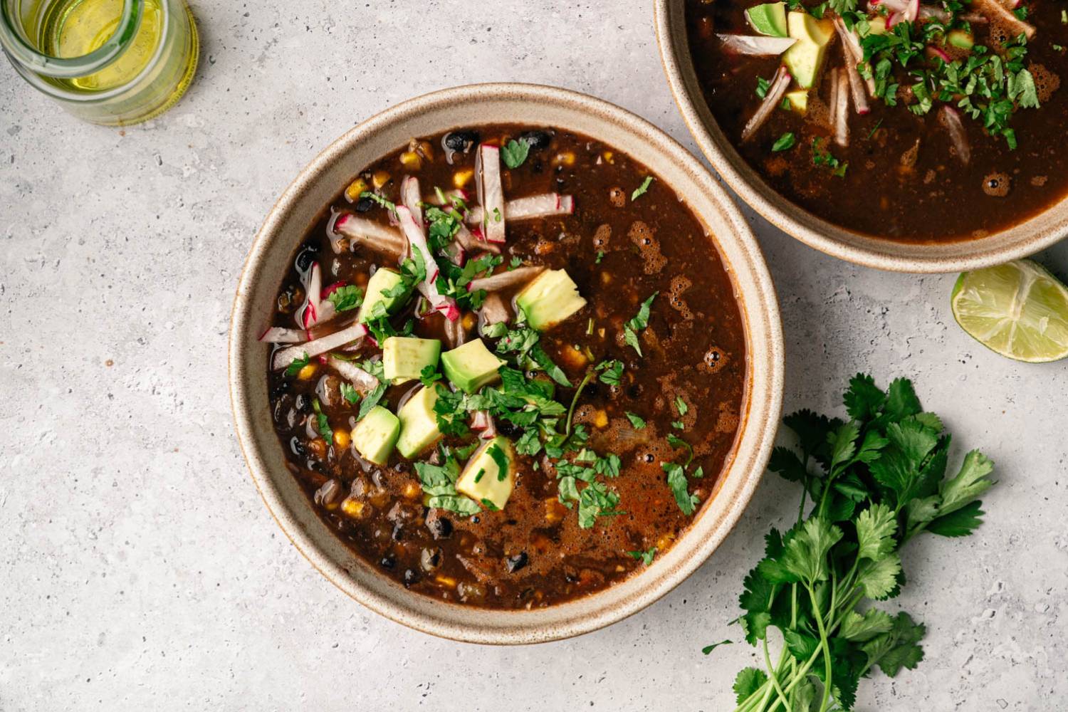 Spicy Black Bean and Corn Soup A bowl of black bean soup topped with avocado, cilantro, and radish strips on a textured gray surface. A jar of oil and fresh lime and cilantro are nearby.