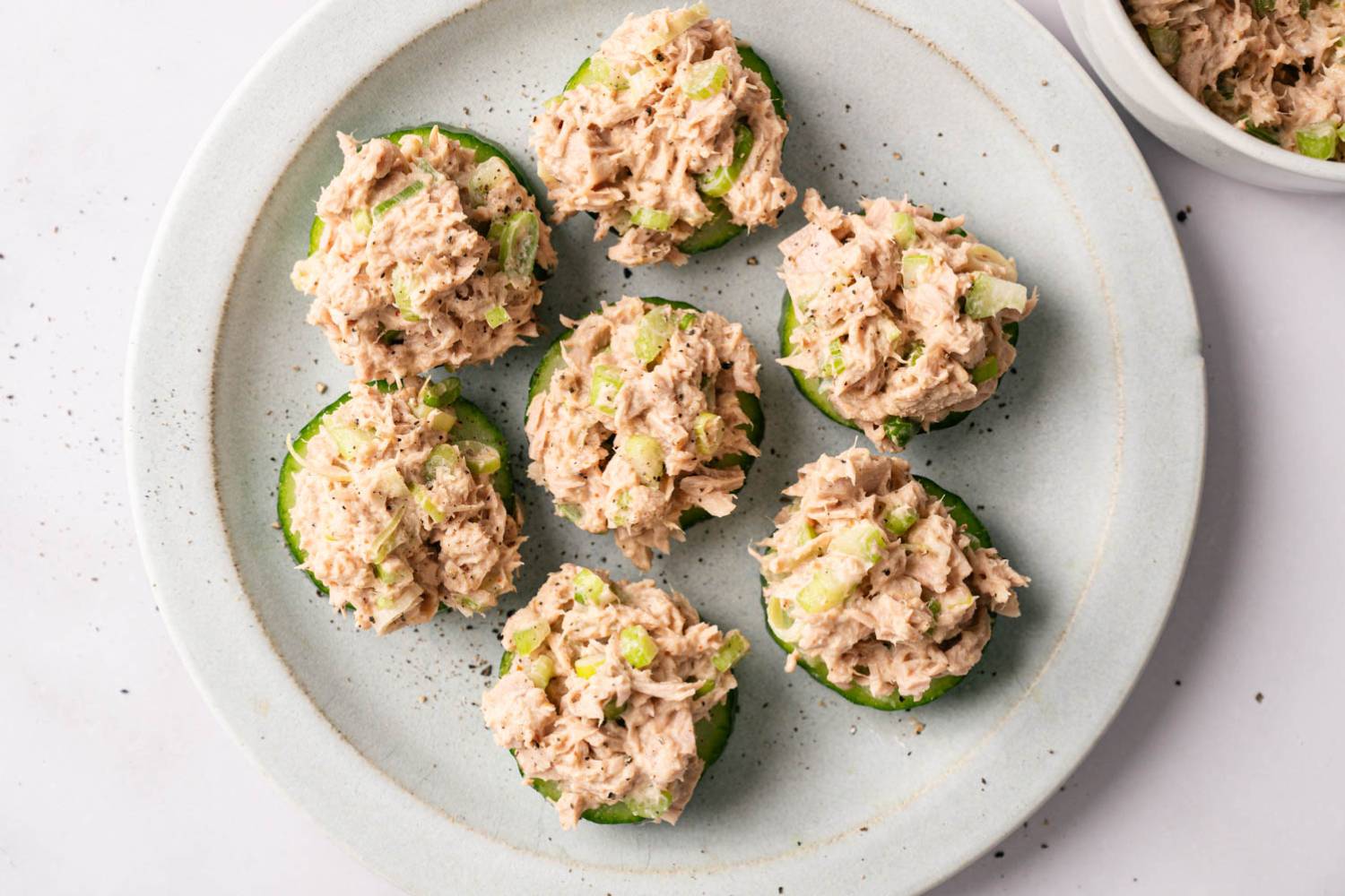 Spicy Tuna Salad Six cucumber slices topped with tuna salad, garnished with celery and pepper, arranged on a white plate. The tone is fresh and appetizing.