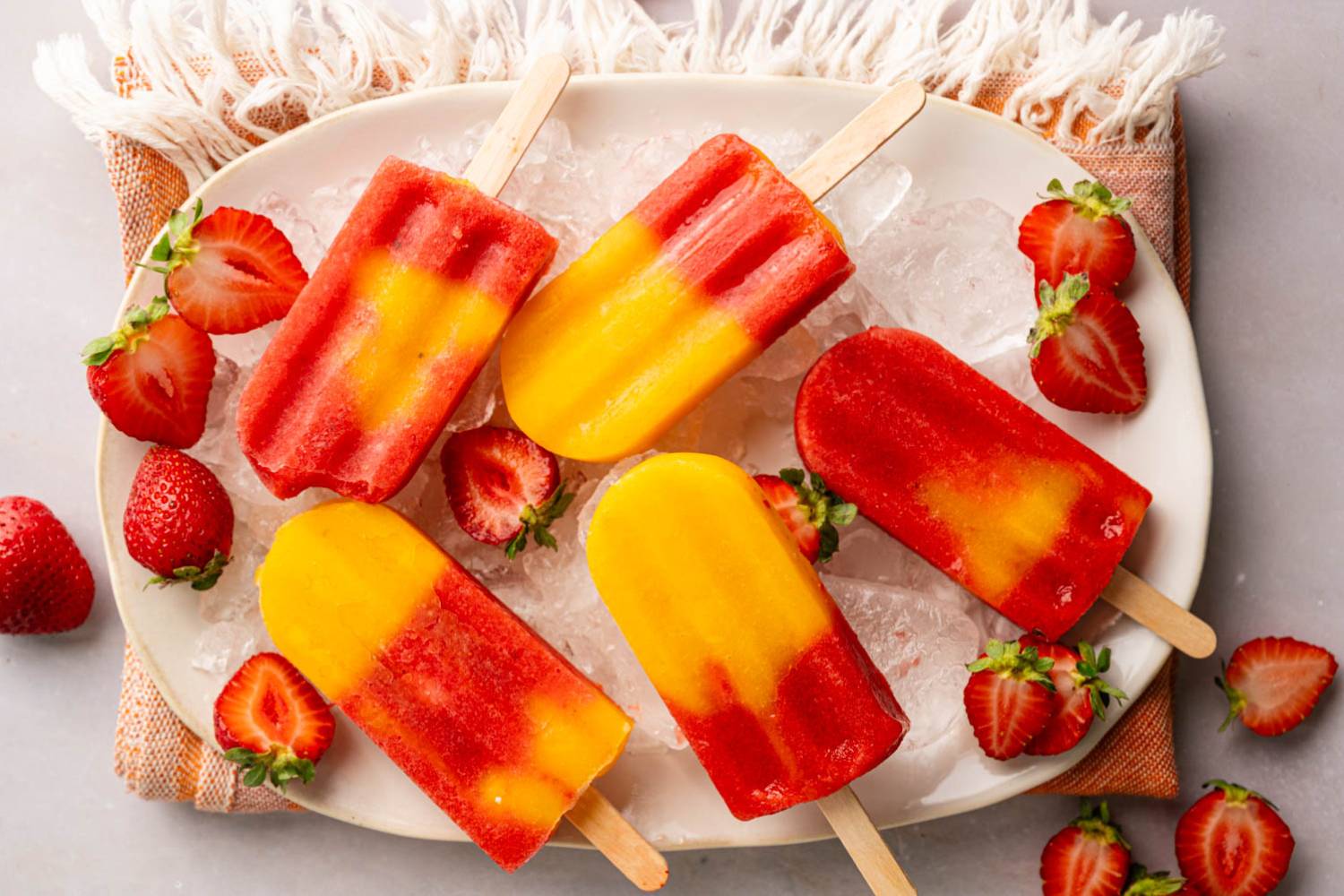 Strawberry Mango Popsicles A platter of layered strawberry mango popsicles on crushed ice, garnished with fresh strawberries for a refreshing summer treat.