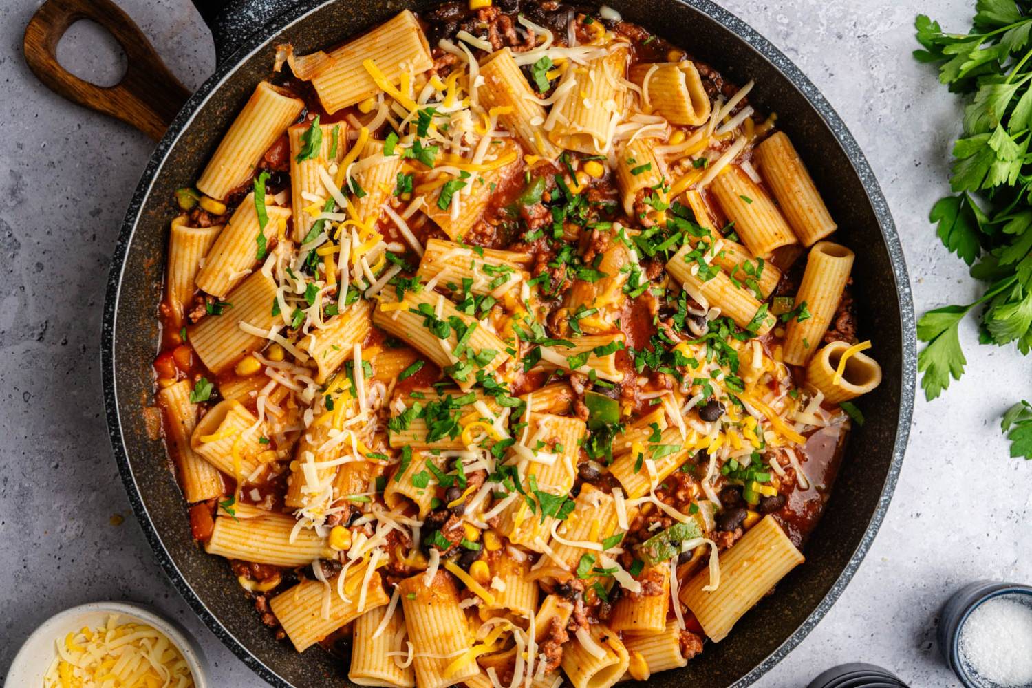 Taco Pasta A pan of rigatoni pasta with tomato sauce, ground meat, corn, and black beans, topped with melted cheese and parsley, on a gray surface.