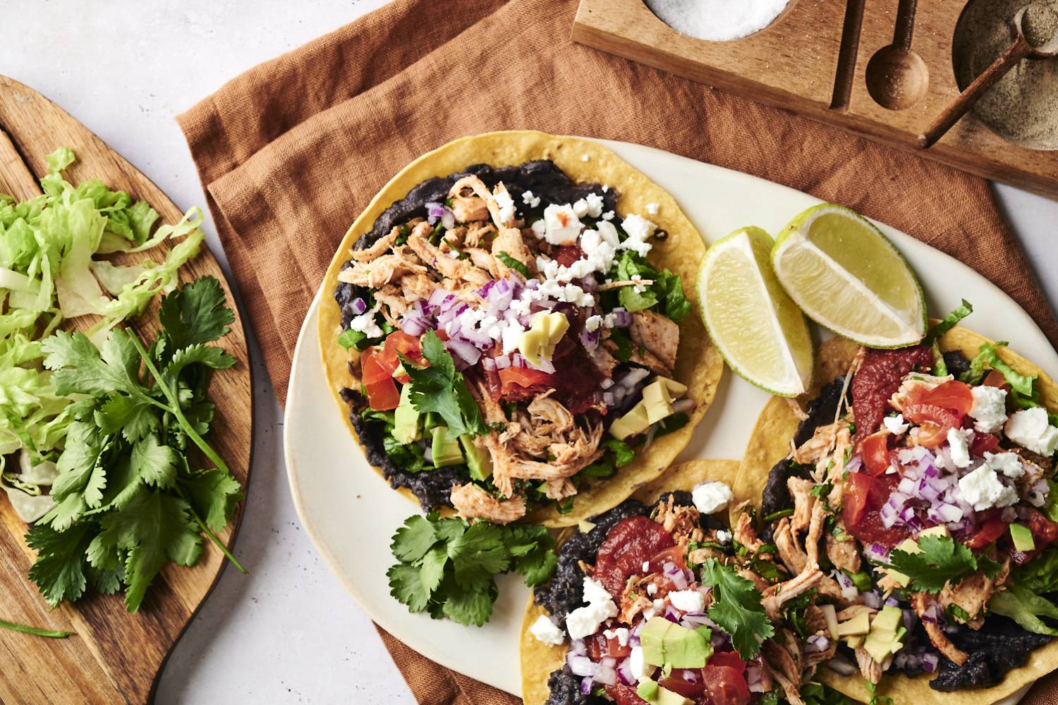 Chicken Tostadas A plate of tostadas, topped with black beans, shredded chicken, fresh lettuce, tomato, red onion, avocado, cilantro, and crumbled cheese. Lime wedges are placed on the side for a fresh squeeze of citrus.