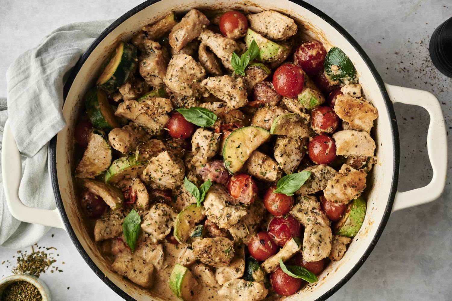 Healthy Tuscan Chicken {Olive Garden Copycat in 30 Minutes!} Top-down view of a creamy Tuscan chicken skillet filled with chunks of seasoned chicken, cherry tomatoes, zucchini slices, and fresh basil in a creamy herb sauce. A gray napkin and small bowl of Italian seasoning rest nearby.
