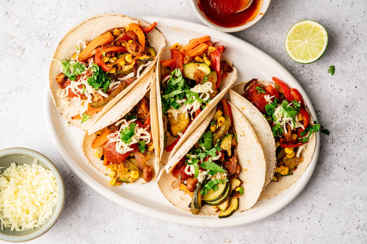 Vegetable Fajitas (Quick and Easy!) A plate with four vibrant vegetable-filled tacos, topped with cilantro and cheese, surrounded by a small bowl of salsa, a lime half, and grated cheese.