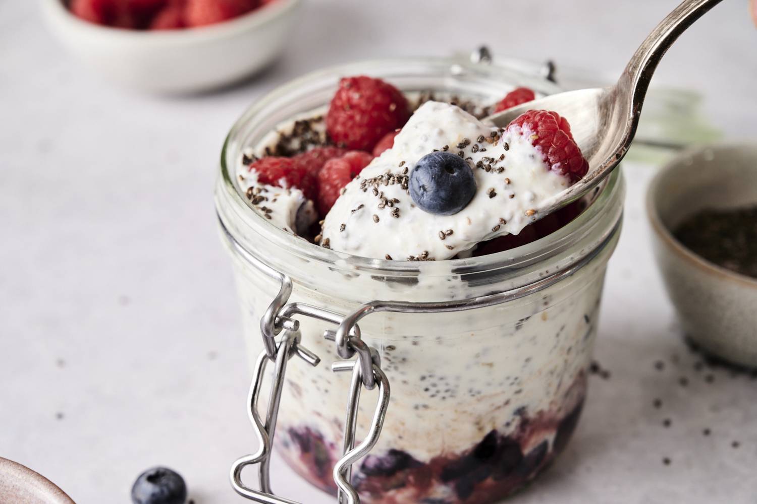 Yogurt Chia Seed Pudding Spoon scooping creamy yogurt chia seed pudding topped with raspberries, blueberries, and chia seeds from a glass jar layered with fruit at the bottom.