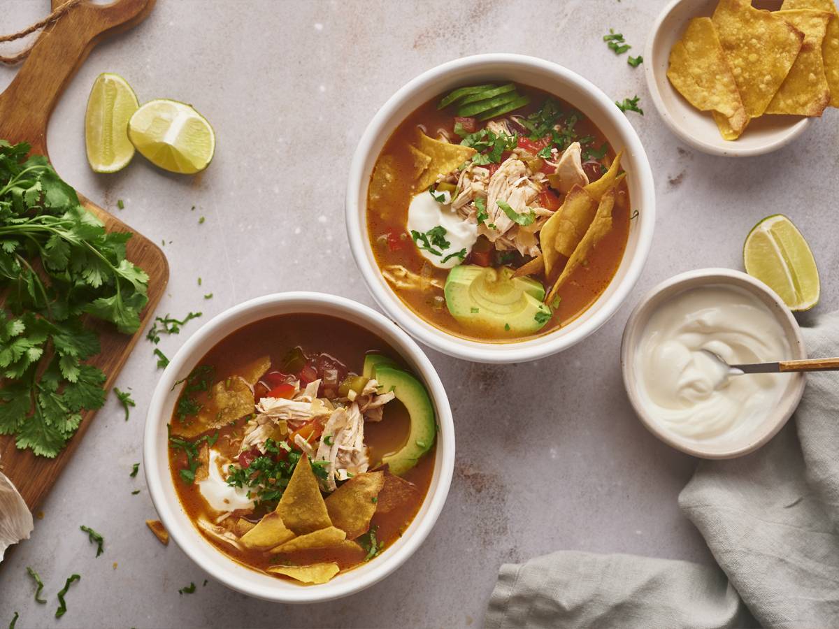 Mexican Dinner Slow Cooker Chicken Fajita Soup in tow bowls with shredded chicken, bell peppers, onions tortilla chips, and avocado.