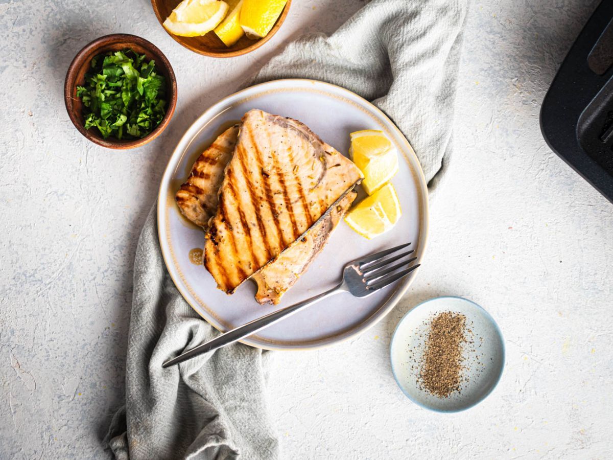 Grilled swordfish on a white plate seen from above with garlic, lemon, and herbs with fresh parsley.