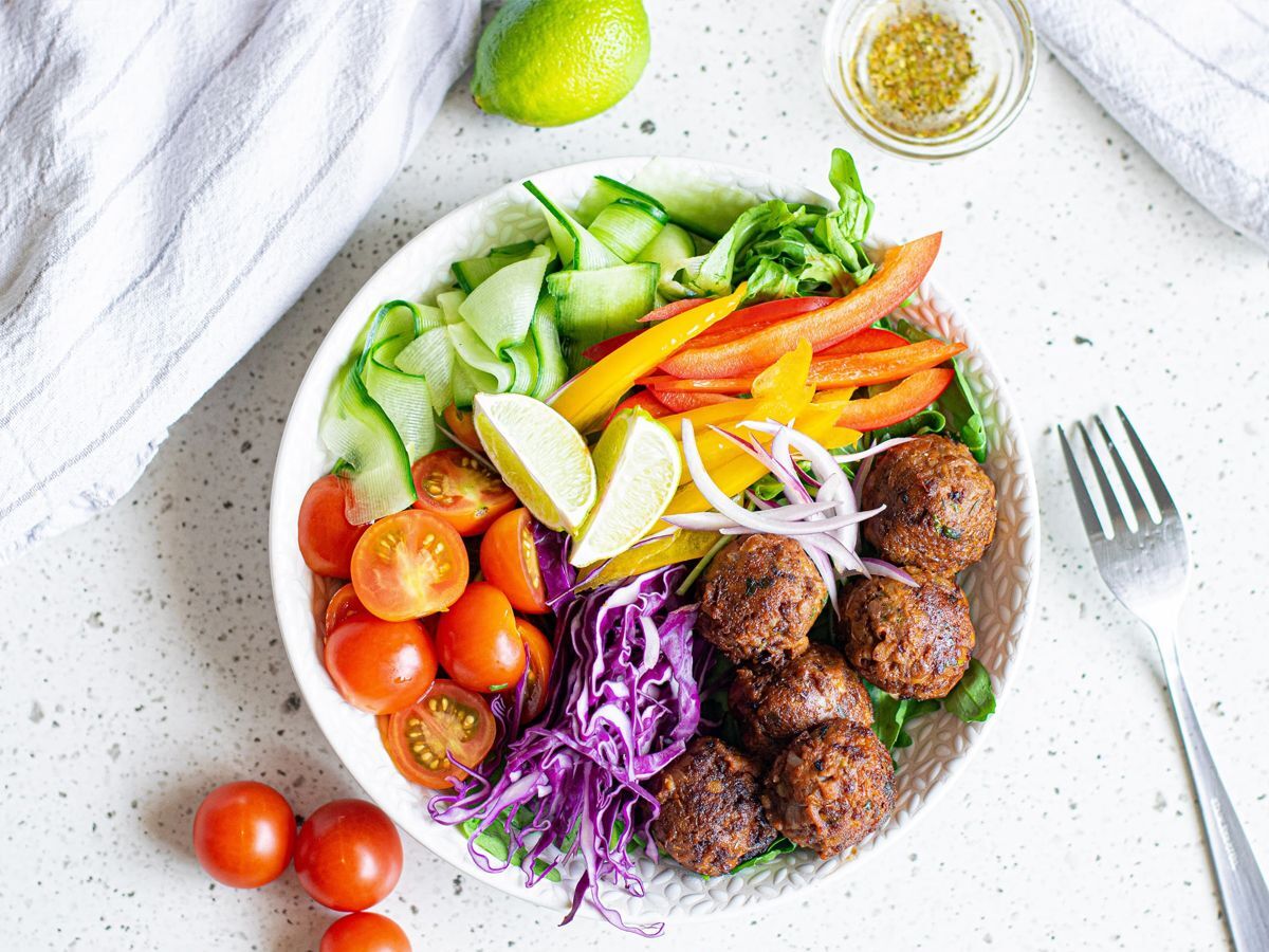 Lentil meatballs served in a bowl with cabbage, peppers, tomatoes, cucumber, red onion, and limes.