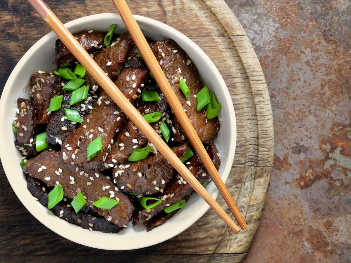 Mongolian Beef in a bowl with chopsticks with scallions and sesame seeds.